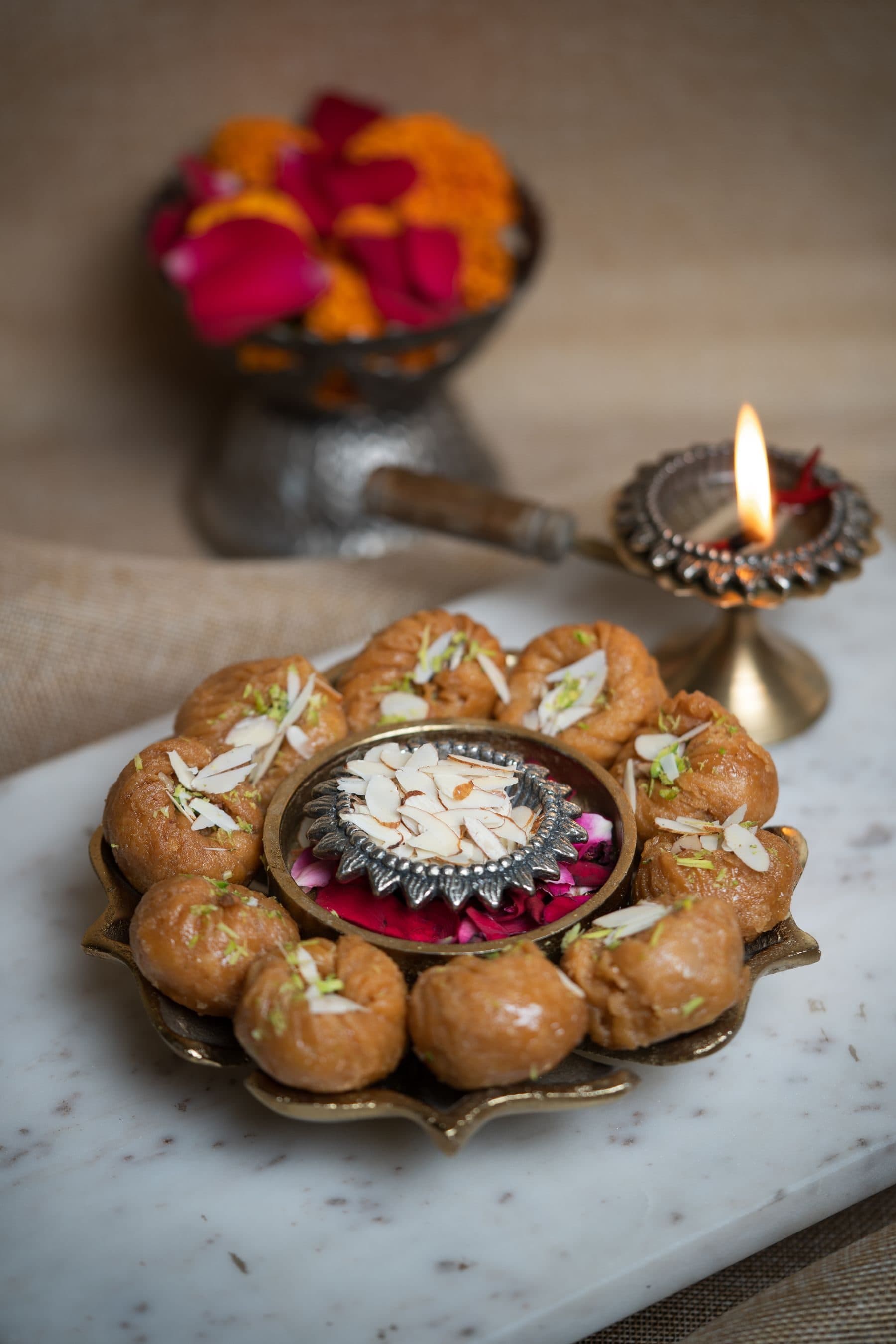 Peda arranged in a ring on a brass tray around a lit diya, with rose petals