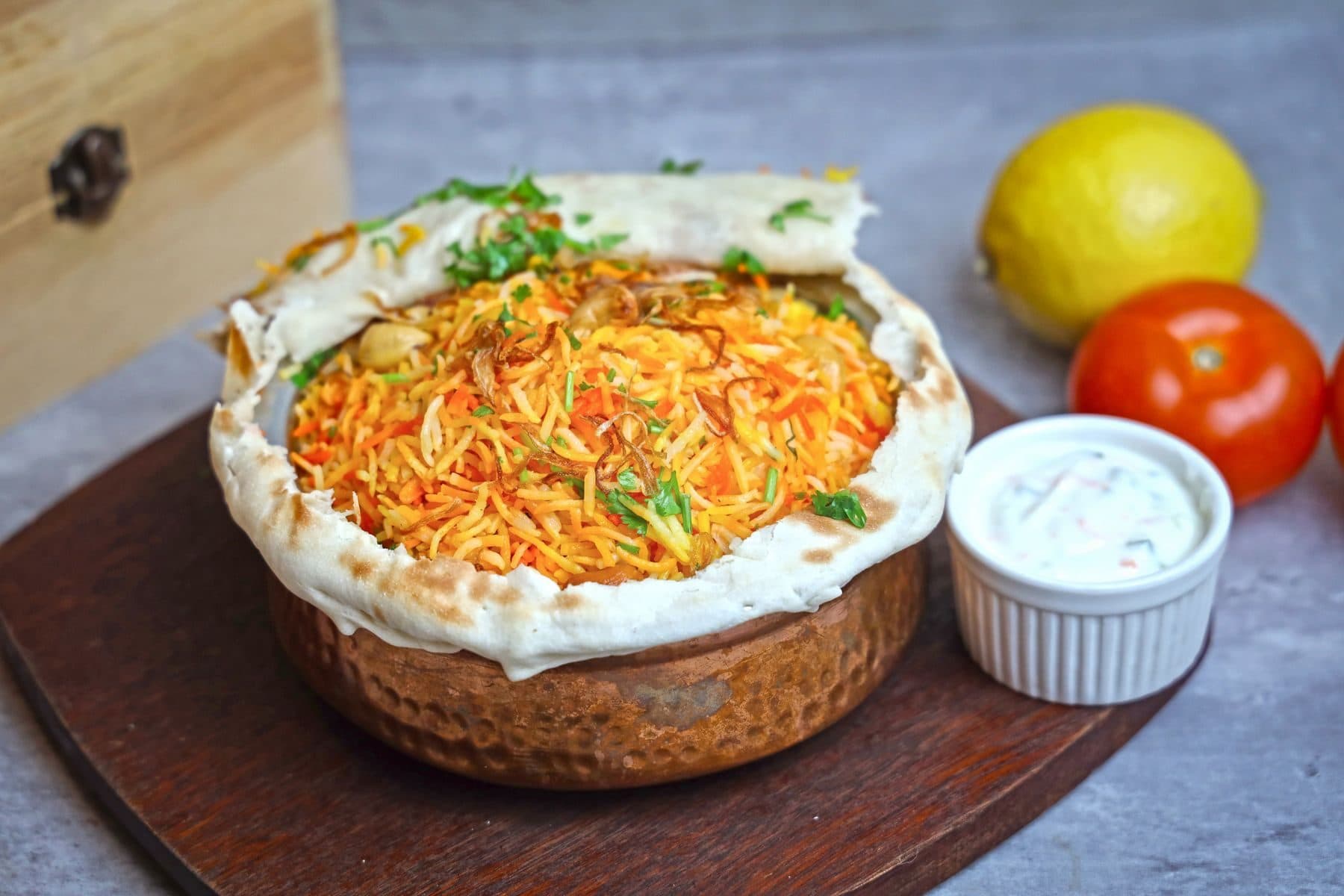 A copper handi of dum biryani sealed with roti, with raita alongside