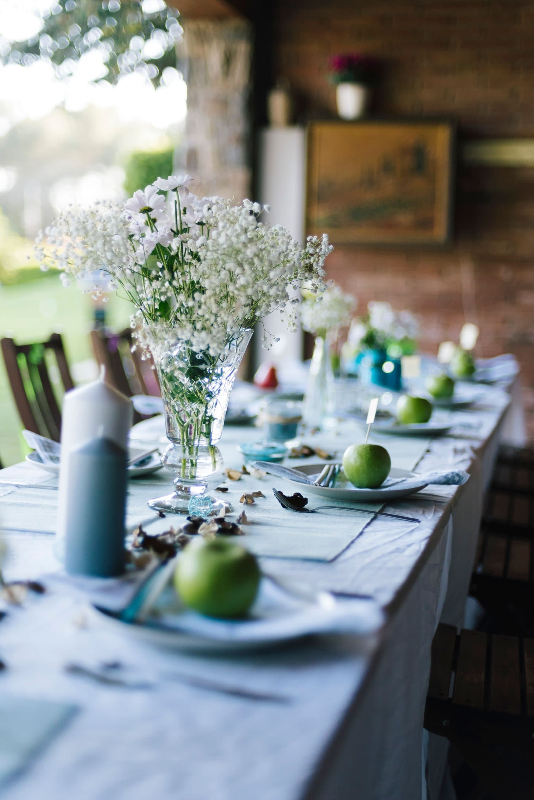 A long lunch table set outdoors with white flowers and green apples