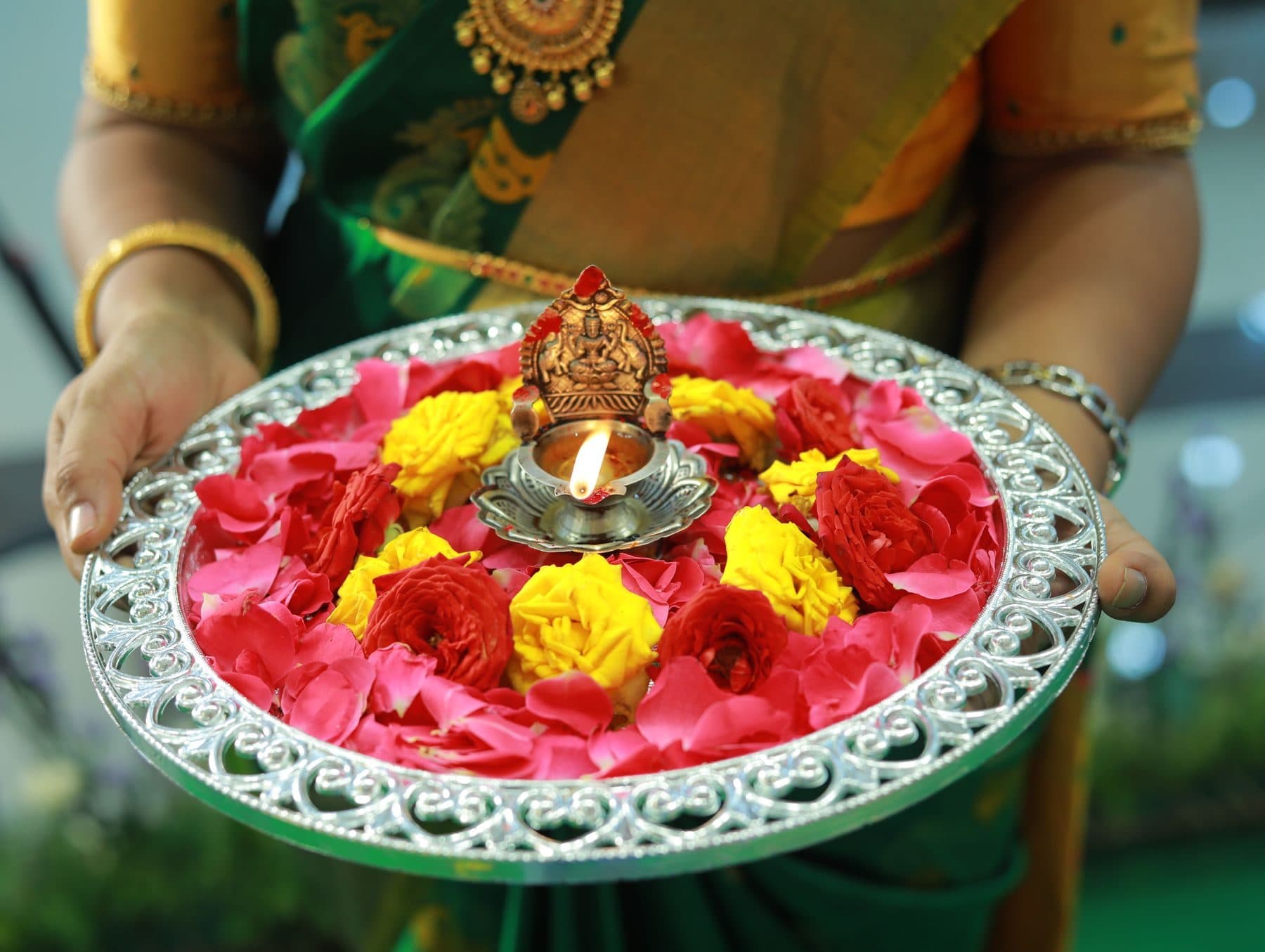 A decorated pooja thali with rose petals, marigolds and a lit lamp held in both hands