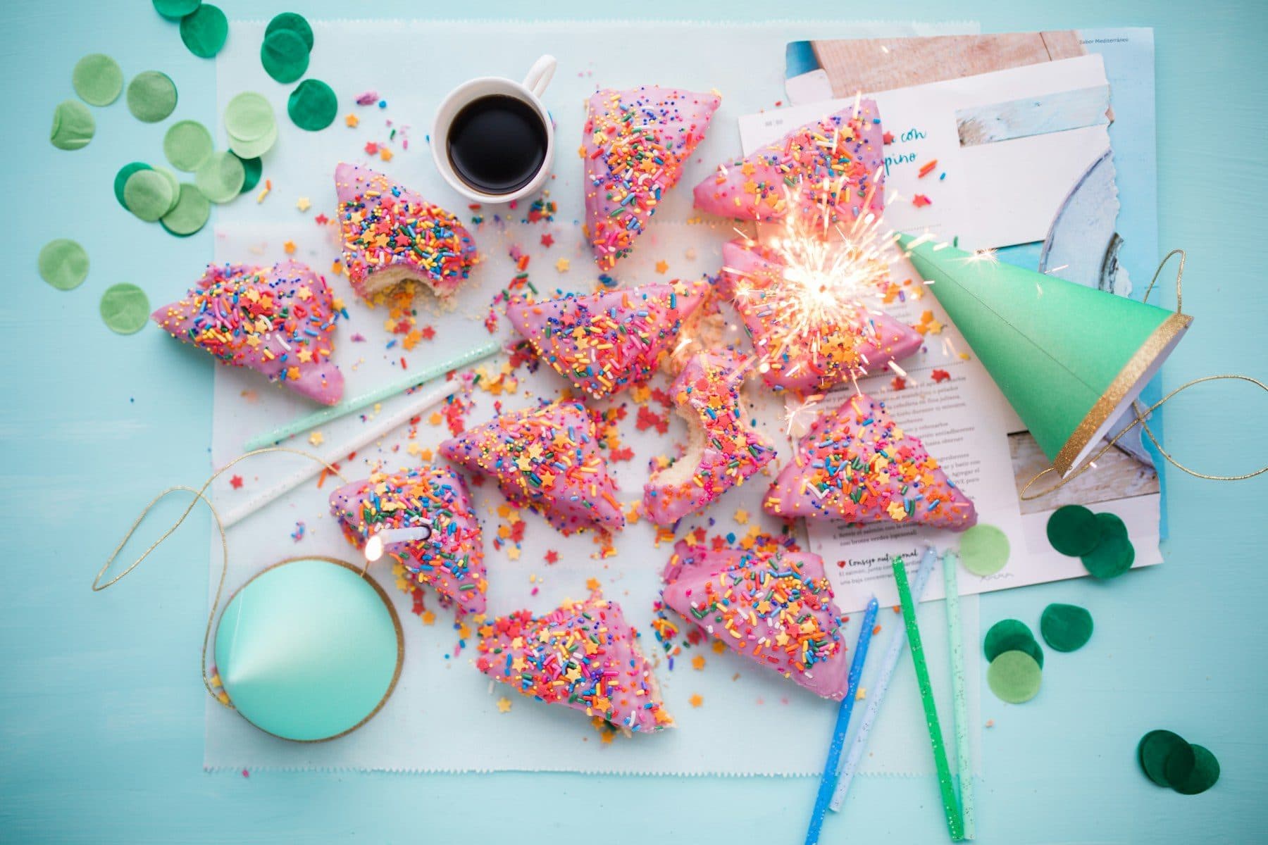 Slices of pink sprinkle cake with a lit sparkler and party hats on a blue table